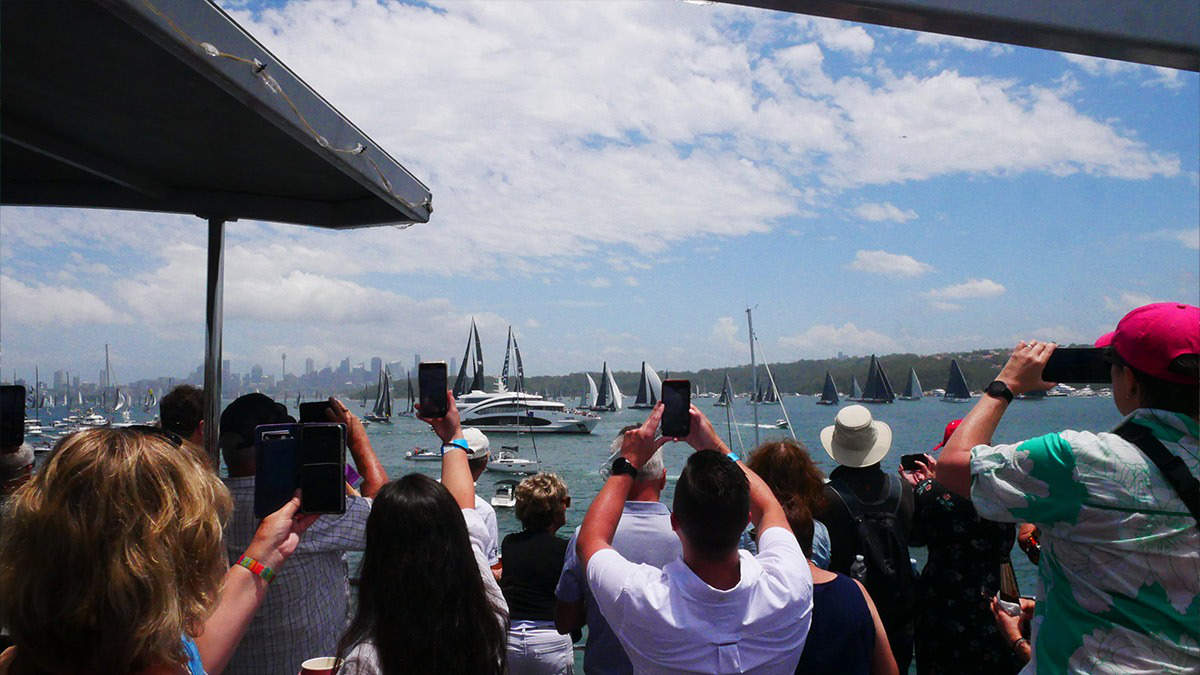 Spectators on a harbour headland watching yachts sail past